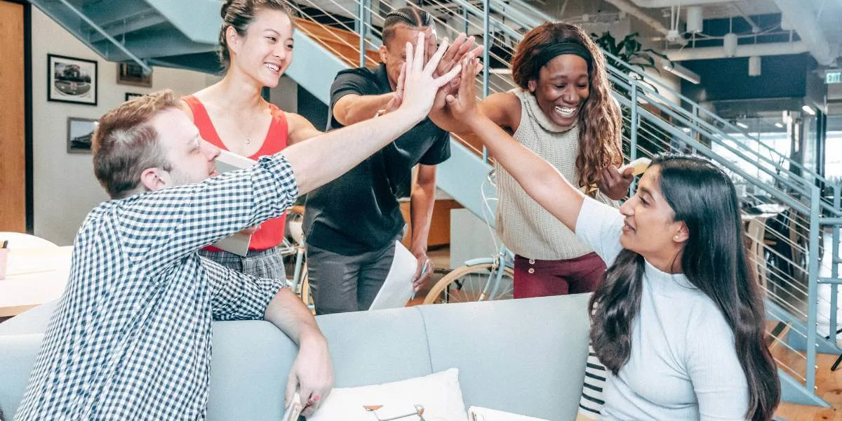 Iloinen tiimi juhlistamassa onnistumista toimistoympäristössä – yhteishenki, yhteistyö ja positiivinen asiakaskokemus keskiössä.